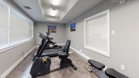 This single-floor fitness room is designed to provide a functional and focused workout environment. The space is finished with neutral gray walls, white baseboards, and durable epoxy-style flooring that offers both practicality and a polished aesthetic. A tray ceiling adds a subtle architectural detail, lending a sense of depth and definition to the room.

Natural light filters in through large windows fitted with white blinds, creating a bright and energizing atmosphere. The room is outfitted with a comprehensive selection of exercise equipment, including a treadmill, elliptical trainer, recumbent stationary bike, and a multi-function cable weight machine, providing options for both cardiovascular and strength training workouts.

Cityscape canvas prints are displayed on the walls, adding a decorative touch and enhancing the overall ambiance of the space. A hand sanitizer dispenser and paper towel holder are mounted near the entry door, reflecting a clean and health-conscious environment. The entry area features carpeted flooring and connects seamlessly to the main workout space, which is equipped with clearly posted informational signage on the door. Overall, the fitness room is well-maintained, thoughtfully organized, and designed to accommodate a well-rounded exercise routine.