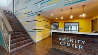 The Circuit Amenity Center is a thoughtfully designed fitness facility that combines energetic aesthetics with comprehensive wellness amenities. Visitors are welcomed by a striking reception area featuring a rustic wood-paneled front desk, bold yellow accent walls, and a dynamic graphic mural of colorful horizontal stripes that extends across the walls and staircase. Warm pendant lighting and wood-look flooring complete the lively, modern entrance. A grand staircase with soaring double-height ceilings adds a dramatic architectural focal point to the lobby.

The facility's fitness areas are organized around motivational wall graphics labeled "energy," "hydrate," and "strength." The well-equipped strength training room features a full suite of weight machines, a cable system, and a large wall mirror, all bathed in natural light from an oversized window. A spacious cardio room lined with windows houses a row of treadmills and elliptical machines, providing an airy environment for endurance workouts.

Two dedicated studio spaces round out the offerings. One room features floor-to-ceiling mirrors, vaulted ceilings, large windows with outdoor views, and open floor space ideal for yoga or dance. A second studio, painted in soft teal, offers balance equipment and large-scale artwork, creating a calm, focused atmosphere for group fitness or stretching sessions. Polished concrete floors and recessed lighting are consistent throughout, tying the facility together with a clean, contemporary finish.
