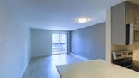 This single-floor space offers a bright and functional layout centered around an open living area. The room features light hardwood-style flooring that extends throughout, complementing the neutral gray and white walls that lend a clean, contemporary aesthetic. A flush-mount ceiling light provides consistent overhead illumination, while a sliding glass door serves as the focal point of the main living area, flooding the space with natural light and providing direct access to an outdoor balcony. The balcony overlooks a treed outdoor setting, offering a pleasant natural view. A baseboard heating unit runs along the exterior wall beneath the sliding door, ensuring efficient warmth throughout the space. A partial countertop or table surface is visible in the foreground, suggesting a defined dining or transitional area nearby. The smooth white ceiling and crisp white baseboards add a polished finish to the overall interior. The open, uncluttered layout allows for versatile furniture arrangements, making the living area adaptable to a variety of needs. Overall, the space presents a well-lit, move-in ready environment with practical features and a calm, neutral ambiance.