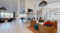 The Cadence Clubhouse in Sugar Hill, Georgia is a thoughtfully designed single-floor community amenity space that blends modern style with a warm, social atmosphere. The entry foyer welcomes guests with large floor-to-ceiling glass doors, white shiplap-style walls, colorful framed artwork, and lush potted greenery set against light tile flooring that extends throughout the entire space.

At the heart of the clubhouse stands a dramatic double-sided fireplace topped with a wall-mounted television, serving as both a functional focal point and a natural divider between activity zones. Bold, oversized pendant light fixtures hang throughout, adding character and depth to the soaring vaulted ceilings.

The lounge area features plush sectional seating, wicker accent chairs, vibrant patterned rugs, and bold botanical wallpaper, creating a relaxed yet lively atmosphere. A dedicated game zone offers a full-size pool table, while a stylish wet bar with a marble-topped counter, rich wood cabinetry, and dramatic tree-motif wallpaper provides an ideal spot for entertaining.

Multiple glass garage-style doors open directly to outdoor patio spaces, flooding the interior with natural light and offering seamless indoor-outdoor connectivity. A casual dining nook with rattan chairs and round tables adds additional gathering space. Together, these elements create a dynamic and inviting community clubhouse designed for connection and leisure.
