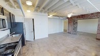 This single-floor unit blends industrial character with modern updates throughout its open, loft-inspired layout. Polished concrete floors extend consistently across all areas, complementing the white-painted exposed beam and joist ceilings that add significant architectural interest and a sense of generous vertical space. Ceiling fans are installed in multiple rooms, enhancing airflow and comfort throughout.

Original brick walls serve as striking accent features in the main living areas, adding raw texture and historic charm that contrast effectively with the clean white walls. Multiple windows allow natural light to filter into the living spaces, brightening the open floor plan considerably.

The kitchen is equipped with stainless steel appliances, including a range, microwave, dishwasher, and refrigerator, alongside dark espresso cabinetry and granite countertops. A dedicated laundry closet with a stacked washer and dryer unit provides added convenience. The bathroom features a full tub-shower combination and continues the polished concrete flooring found throughout the unit. A bedroom includes a closet and ceiling fan. The overall ambiance is clean, functional, and distinctly industrial in character.