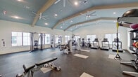 This fitness center is a standout facility designed to support a full range of workout routines. The space is defined by its striking vaulted ceiling, painted in a cool blue-gray tone and accented by exposed beam details, which lends the room an open, airy atmosphere. Multiple large ceiling fans are mounted throughout, ensuring consistent airflow across the entire floor. Recessed lighting supplements the abundant natural light that pours in through numerous oversized windows positioned along multiple walls, offering views of the surrounding exterior architecture.

The dark rubber flooring provides a durable, high-performance surface throughout, with yoga and stretching mats placed strategically across the floor. The strength training area is well-appointed, featuring a full dumbbell rack, multiple cable and weight machines including Hoist-brand multi-functional units, a power rack with bench press setup, medicine balls, and resistance training accessories. Floor-to-ceiling mirrors line one wall, amplifying the sense of space and allowing users to monitor their form.

The cardio section is equally comprehensive, housing a row of treadmills, StairMaster steppers, and elliptical machines — all positioned near windows for an engaging workout environment. The overall layout is thoughtfully organized, providing ample open floor space between equipment zones for stretching, functional training, and unobstructed movement throughout the facility.