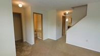 This two-story residence offers a practical and straightforward layout designed for comfortable everyday living. The main floor features a carpeted living area that provides a warm and inviting atmosphere, with neutral white walls that create a bright, open feel throughout the space. A full bathroom is conveniently located on the main level, outfitted with a vanity, cabinetry, and standard fixtures with warm wood-toned trim framing the doorway. Adjacent to the living area, a hallway leads toward the kitchen, which is equipped with cabinetry and essential appliances, offering a functional workspace for meal preparation. A staircase with a wood railing provides access to the upper floor, suggesting additional living or sleeping quarters above. Ceiling-mounted light fixtures illuminate the main floor corridors and common areas. The space features consistent neutral tones, carpeted flooring in the main living zone, and hard flooring in the bathroom and transitional areas. Visible HVAC vents indicate climate control throughout. Overall, the home presents a clean, no-frills interior well-suited for those seeking a straightforward, multi-level living arrangement.