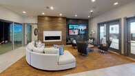 This thoughtfully designed single-floor amenity space blends contemporary style with functional comfort across several distinct zones. The lounge area serves as a central gathering hub, anchored by a sleek linear fireplace set within a striking wood-slat accent wall. Adjacent to it, a textured dark feature wall hosts multiple large flat-screen televisions, making it an ideal spot for entertainment. Comfortable curved sectional sofas and modern swivel chairs are arranged throughout, resting on warm patterned area rugs atop light tile flooring, all beneath recessed ceiling lighting that creates a welcoming ambiance.

The kitchen and dining area is equally impressive, showcasing dark cabinetry with a white subway tile backsplash, stainless steel appliances, and a generous white quartz island with a solid wood base. Pendant lights hang above the island, which is surrounded by upholstered counter-height chairs in a rich terracotta tone. French doors along the exterior wall flood the space with natural light and provide access to an outdoor area.

A separate games and work zone, visible in the floorplan view, features a dartboard, communal work tables, and additional seating, offering a versatile space for both recreation and productivity. The overall design is cohesive and modern, with a warm material palette of wood, stone, and metal finishes throughout.