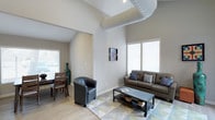 This thoughtfully designed single-floor space makes a striking first impression with its soaring vaulted ceilings that lend an expansive, airy quality to the interior. The living area serves as the heart of the space, anchored by a plush leather sofa and a coordinating accent chair arranged around a sleek, industrial-style coffee table. A geometric patterned area rug in soft, muted tones defines the seating arrangement and adds a contemporary touch to the light hardwood flooring beneath.

Natural light pours in generously through multiple windows fitted with clean white horizontal blinds, brightening the neutral gray walls and creating a warm, welcoming ambiance throughout. A sculptural pendant light fixture descends elegantly from the peak of the vaulted ceiling, serving as both a functional and decorative focal point.

A dedicated dining area is visible just beyond the living space, featuring a wooden dining table and chairs, seamlessly integrated into the open-concept floor plan. The layout promotes a smooth flow between living and dining areas, making the space feel connected and versatile. Wall-mounted artwork adds a personal touch, while the overall design palette — neutral tones, clean lines, and modern furnishings — creates a cohesive, contemporary aesthetic throughout.