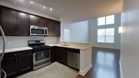 This two-floor space offers a well-designed interior with a contemporary aesthetic throughout. The kitchen is a central highlight, featuring rich dark-toned cabinetry that contrasts elegantly with light countertops. Stainless steel appliances, including a gas range, built-in microwave, and dishwasher, provide a sleek, modern finish. Track lighting overhead ensures the kitchen is well-illuminated, while tile flooring defines the cooking area. A peninsula with a built-in sink and breakfast bar provides additional workspace and serves as a natural transition into the adjacent living area. The living space features warm hardwood flooring and benefits from generous natural light streaming through large windows, creating a bright and welcoming atmosphere. The open-concept layout between the kitchen and living area promotes a fluid, connected feel ideal for both everyday living and entertaining. The combination of modern finishes, thoughtful design, and natural light gives this two-story space a clean, contemporary character throughout.