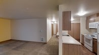 This single-floor space offers a practical and functional layout designed for comfortable everyday living. The open-concept design connects the kitchen and living area seamlessly, creating a cohesive and easy-to-navigate floor plan. The kitchen features warm wood-toned cabinetry, a clean white countertop, and a stainless steel sink, providing a well-equipped and organized workspace. A partial wall with a pass-through counter separates the kitchen from the living area while maintaining an open feel. The living area is finished with plush wall-to-wall carpeting in a neutral beige tone, contributing to a warm and inviting atmosphere. The walls are painted in a soft, neutral white that reflects light evenly throughout the space. A hallway extends from the main living area, lined with doors leading to additional rooms, suggesting a thoughtful separation of private and communal spaces. Built-in HVAC venting is present in the living area, ensuring climate comfort year-round. Ceiling-mounted lighting provides consistent illumination throughout. Overall, the space presents a clean, understated aesthetic with functional finishes suited to a wide range of lifestyles.