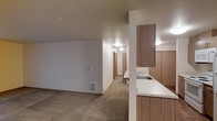 This single-floor space offers a practical and functional layout designed for comfortable everyday living. The open-concept design connects the kitchen and living area seamlessly, creating a cohesive and easy-to-navigate floor plan. The kitchen features warm wood-toned cabinetry, a clean white countertop, and a stainless steel sink, providing a well-equipped and organized workspace. A partial wall with a pass-through counter separates the kitchen from the living area while maintaining an open feel. The living area is finished with plush wall-to-wall carpeting in a neutral beige tone, contributing to a warm and inviting atmosphere. The walls are painted in a soft, neutral white that reflects light evenly throughout the space. A hallway extends from the main living area, lined with doors leading to additional rooms, suggesting a thoughtful separation of private and communal spaces. Built-in HVAC venting is present in the living area, ensuring climate comfort year-round. Ceiling-mounted lighting provides consistent illumination throughout. Overall, the space presents a clean, understated aesthetic with functional finishes suited to a wide range of lifestyles.