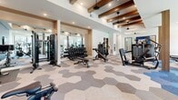 Located at 600 Winners Cir in Rowlett, Texas, this well-appointed fitness center offers an expansive, single-floor layout designed to accommodate a wide range of workout needs. The interior is defined by its striking hexagonal-patterned carpet in neutral tones of blue, gray, and beige, which adds a contemporary visual identity to the space. Decorative wood beam accents line the ceiling alongside recessed lighting and ceiling fans, creating a warm yet modern ambiance throughout.

The main workout floor is generously equipped with treadmills, ellipticals, stationary bikes, cable machines, a dumbbell rack, and various strength-training benches and weight machines. Large windows along the perimeter flood the space with natural light and offer views of the surrounding outdoor areas. A bold blue accent wall serves as a focal point for a dedicated cycling station, complete with a spin bike and wall-mounted television for guided workouts.

A separate enclosed studio area, accessible through glass-panel doors, provides space for functional training, featuring additional equipment and ample room for movement-based exercises. The facility also includes a water station and built-in storage cabinetry, adding practical convenience. Full-length mirrors in the strength training zone enhance the sense of openness and allow users to monitor their form effectively.