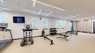 This fitness center offers a spacious, single-floor exercise room designed to support a full range of workout routines. The interior is finished with clean white walls and durable speckled flooring, creating a bright and hygienic environment well-suited for physical activity. A striking geometric pattern of linear LED lighting spans the ceiling, providing even, high-quality illumination throughout the space. Multiple windows fitted with horizontal blinds line the walls, allowing natural light to filter in while maintaining privacy.

The cardio zone is equipped with two treadmills, an elliptical trainer, and a rowing machine, offering users a variety of aerobic workout options. A wall-mounted flat-screen television is positioned for easy viewing during cardio sessions. The strength training area includes a multi-station cable machine, a power rack, adjustable weight benches, and a well-stocked dumbbell rack, accommodating both beginner and advanced lifters. A large floor-to-ceiling mirror along one wall enhances the sense of openness and assists users with form and technique.

Additional accessories such as stability balls are stored in a dedicated corner area. Floor-to-ceiling glass doors provide a visual connection to an adjacent lounge area, adding to the open and connected feel of the space. The fitness center is thoughtfully organized to maximize usability while maintaining a clean, modern aesthetic.