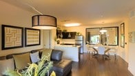 This thoughtfully designed single-floor space showcases an open-concept layout that effortlessly connects the living, kitchen, and dining areas into one cohesive and inviting environment. Rich dark hardwood flooring runs throughout, anchoring the space with warmth and continuity.

The living area is highlighted by a striking oversized drum pendant light and a sleek dark leather sectional sofa, creating a comfortable and contemporary focal point. A bold striped framed artwork adds a pop of color and personality to the walls.

The kitchen is both functional and stylish, featuring dark cabinetry with contrasting light countertops, stainless steel appliances including a full-size refrigerator, and elegant teardrop pendant lights that hang above the peninsula. The peninsula itself provides additional workspace and casual seating opportunities.

The dining area flows naturally from the kitchen, bathed in natural light from a large window that brings the outdoors in. A modern dining table is surrounded by sleek white chairs, illuminated by a contemporary pendant light fixture overhead.

Throughout the space, lush indoor greenery adds life and vibrancy, complementing the modern aesthetic. The overall ambiance is warm, polished, and welcoming, making excellent use of every corner of the open floor plan.