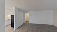 This single-floor unit offers a clean, functional layout with a neutral color palette throughout. The living area is open and spacious, featuring durable wood-look laminate flooring in a warm gray tone, crisp white walls, and a textured ceiling. A sliding glass door fitted with vertical blinds allows natural light into the main living space, while a doorway connects seamlessly to the rest of the unit.

The galley-style kitchen is efficiently designed with white cabinetry on both sides, light quartz-style countertops, a double stainless steel sink, and black appliances including a full-size refrigerator. Overhead lighting provides consistent illumination throughout the workspace.

The bedroom continues the wood-look flooring and maintains the same neutral aesthetic, offering a calm and versatile space. A ceiling fan with light fixture is present in the adjoining hallway area, which also features a floor-mounted HVAC vent for climate control.

The bathroom includes a vanity with a light-up mirror, light-toned countertops, and a full tub and shower combination with white tile surround and a chrome towel bar. The flooring transitions into the same wood-look planks found throughout the unit, contributing to a cohesive overall appearance.