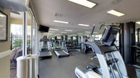 This single-floor fitness center offers a thoughtfully organized workout environment designed to accommodate a wide range of exercise routines. The space is outfitted with multiple treadmills and elliptical machines, providing ample cardio options for users at any fitness level. A dedicated free weights section features a full dumbbell rack, making strength training accessible and convenient. Floor-to-ceiling mirrors line the walls, visually expanding the space and allowing users to monitor their form during workouts. Overhead fluorescent lighting ensures the room is well-lit throughout, complemented by large windows along one side that invite natural light and views of the outdoors. The flooring consists of durable rubber gym matting, offering both safety and comfort underfoot. A wall-mounted flat-screen television is positioned for easy viewing from the cardio equipment, enhancing the workout experience. The textured ceiling adds a classic finish to the space. Overall, the fitness center presents a clean, functional, and well-maintained environment equipped to support a comprehensive fitness regimen.