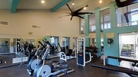 This fitness center is designed to offer a comprehensive workout experience within a spacious, airy environment. The room immediately impresses with its soaring ceilings, accented by distinctive teal-painted beams and columns that lend the space a vibrant, energizing character. A large ceiling fan hangs prominently overhead, promoting airflow throughout the workout area and contributing to a comfortable atmosphere during exercise.

The flooring is a dark, durable material suited for high-traffic fitness use, providing both safety and functionality. Full-length mirrors line the walls, visually expanding the space and allowing users to monitor their form during workouts. Natural light filters in through multiple windows, further brightening the interior.

The equipment layout is thoughtfully organized, featuring a row of cardio machines — including ellipticals and treadmills — positioned along one side of the room. A cable and weight machine occupies the center, while a well-stocked dumbbell rack lines the opposite wall, catering to free weight training. Recessed lighting supplements the natural light, ensuring the space remains well-lit at all hours. Overall, this fitness center combines functional design with an inviting aesthetic, making it a motivating environment for a wide range of exercise routines.