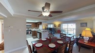 This single-floor residence features an open-concept layout that seamlessly connects the kitchen, dining area, and living room, creating a cohesive and functional living environment. The kitchen is well-appointed with dark wood cabinetry, granite countertops, and stainless steel appliances including a range, microwave, and dishwasher. A peninsula with seating provides additional workspace and casual dining options. Adjacent to the kitchen, the dining area accommodates a substantial dark wood table with seating for six, set beneath a five-blade ceiling fan with integrated lighting. Crown molding and a textured ceiling add architectural detail throughout the space. The living room is anchored by gleaming light-toned tile flooring and features large sliding glass doors that flood the room with natural light and open directly onto a balcony with expansive city views. A leather sofa, accent chair, and framed artwork complete the living area's furnishings. The balcony extends the living space outdoors, offering a comfortable setting to enjoy the surrounding urban panorama. The overall ambiance is bright, clean, and well-maintained.