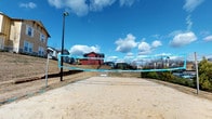 This outdoor sand volleyball court is a well-appointed recreational amenity situated within a residential community. The court features a regulation-height net with bright blue accent banding, clearly defined boundary lines, and a level, fine-sand playing surface designed for comfortable athletic play. A tall, dedicated light pole stands adjacent to the court, enabling use during evening hours and extending recreational opportunities beyond daylight.

The court is open-air, offering expansive views of the surrounding neighborhood, including multi-story residential buildings and mature trees that provide a pleasant backdrop. The open sky above floods the space with natural light, creating an inviting atmosphere for both casual and competitive play.

The surrounding grounds include landscaped green areas, walking paths, and seating elements nearby, making the space suitable for spectators and community gatherings. Additional functional infrastructure is visible in the vicinity, including utility access points and building entrances with classic craftsman-style exterior details, reflecting the broader architectural character of the community. The overall setting is designed to encourage outdoor activity and community engagement, offering a well-maintained and accessible recreational space for residents.