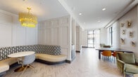 This elegantly designed single-floor space functions as a stylish communal lounge, combining refined architectural details with carefully curated furnishings to create a welcoming and visually dynamic environment. Throughout the space, light-toned herringbone-patterned flooring establishes a cohesive foundation, complemented by crisp white wainscoting and raised panel wall treatments that lend a classic yet contemporary character.

The layout is thoughtfully organized into distinct zones. A central lounge area features a built-in curved banquette with a patterned back cushion, positioned around a round table beneath a striking gold tiered chandelier. An intimate nook tucked into the corner showcases a mustard-yellow L-shaped bench set against a richly textured, oxidized wall panel, illuminated by elegant marble-accented wall sconces. Decorative throw pillows and a coordinating yellow side table complete the vignette.

Toward the street-facing end of the space, floor-to-ceiling black-framed windows allow abundant natural light to pour in, offering open views of the surrounding streetscape. Sheer white drapes soften the light. This area accommodates a mix of green velvet lounge chairs, a dark communal table with vibrant orange seating, and a brass drum coffee table. A cluster of sculptural gold wall fixtures adds an artistic focal point. The overall ambiance is polished, warm, and inviting.