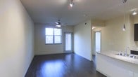 This single-floor space features an open layout with dark hardwood flooring that runs throughout the main living area, lending a warm and polished aesthetic. The living area is well-lit, benefiting from multiple windows fitted with blinds that allow for natural light control. A ceiling fan with an integrated light fixture is centrally positioned, while track lighting on the ceiling provides additional illumination. The neutral, light-toned walls create a clean and versatile backdrop, complementing the darker flooring. A partial view into an adjacent area reveals a countertop with a light granite or stone surface, suggesting a nearby kitchen space. A doorway visible toward the rear of the unit leads to what appears to be a secondary room or hallway, hinting at a connected layout beyond the main living space. The overall ambiance is modern and understated, with practical finishes suited to everyday living. The combination of natural light, contemporary lighting fixtures, and quality flooring gives the space a bright and functional character throughout.