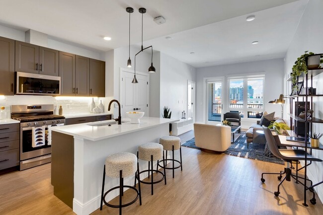 Modern open-plan kitchen and living area with sleek cabinetry, a chic island with bar stools, and large windows offering vibrant city views. - The Fitzgerald