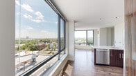 Located on the ninth floor at 199 N Harvard Street in Boston's Allston neighborhood, Unit N906 is a thoughtfully designed one-bedroom, one-bathroom residence that makes a strong impression with its modern aesthetic and expansive city views. The open-concept living and kitchen area is anchored by floor-to-ceiling windows that wrap the space in natural light while offering sweeping panoramic views of the Boston skyline and surrounding neighborhood treetops. Rich hardwood flooring flows throughout, complementing the contemporary design. The kitchen is a standout feature, showcasing high-gloss, wood-grain cabinetry in a deep walnut tone, a white quartz peninsula with an integrated sink, white subway tile backsplash, and a full suite of stainless steel appliances. The bedroom offers a clean, bright retreat with access to a dedicated walk-in closet equipped with built-in wire shelving for organized storage. A hallway corridor with additional window views connects the living space to the private quarters. The bathroom features classic white subway tile surrounding a full soaking tub and shower combination. The entrance foyer provides a natural transition into the home. Clean white walls throughout create a neutral canvas, while the unit's elevated position ensures an unobstructed and light-filled living experience.