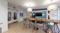 This well-appointed communal space combines contemporary design with practical amenities across an open, single-level layout. The centerpiece is a striking staircase with dark wood treads and sleek stainless steel cable railings, rising alongside an elevated lounge area fitted with a long, cushioned green sectional sofa accented by colorful decorative pillows. Cylindrical metallic stools provide casual seating nearby, while landscape artwork adorns the crisp white walls above.

Light tile flooring flows throughout, unifying the various functional zones. A café-style bar area features a warm wood communal table with teal upholstered stools beneath a large pendant light, paired with a wall-mounted flat-screen TV set against a bold teal accent wall, along with a beverage station and under-counter refrigeration. An adjacent business center provides a built-in desk with multiple workstations and ergonomic chairs, benefiting from natural light through large glass entry doors that offer views of exterior greenery. Restroom facilities are conveniently accessible nearby. The overall ambiance is bright, modern, and welcoming, with a thoughtful blend of social and productive spaces.