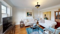 This thoughtfully designed single-floor residence offers a comfortable and cohesive living experience across several well-defined spaces, all unified by warm hardwood flooring and a neutral, cream-toned color palette throughout.

The living room serves as the heart of the home, anchored by a classic white fireplace mantle and adorned with elegant crown molding. A decorative pendant light adds character overhead, while generous windows allow natural light to fill the room. An adjoining media area features a wide bay window with urban views, providing an inviting space to relax.

The formal dining room is highlighted by a stunning ornate chandelier with crystal and amber accents, complemented by multiple windows that bathe the space in natural light. Crown molding continues here, reinforcing the home's traditional architectural details.

The kitchen is equipped with warm honey-toned wood cabinetry and stone-patterned granite countertops, offering ample workspace and storage. The bedroom is a serene retreat, featuring an elegant wrought iron bed frame, natural light from a single window, and direct access to the hallway. The bathroom is finished with classic white subway tile wainscoting, a framed mirror, and chrome fixtures for a clean, timeless look. A dedicated storage closet with built-in shelving adds practical functionality to round out this well-organized home.