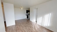 This single-floor space features an open-concept layout that seamlessly connects the living area to the kitchen. The living area is finished with light hardwood-style flooring that reflects natural light streaming in from nearby windows, contributing to a bright and airy atmosphere. White walls throughout the space enhance the sense of openness and provide a clean, neutral backdrop.

A ceiling fan is centrally mounted, offering both lighting and air circulation to the main living area. The kitchen, visible from the living space, is compact yet functional, featuring dark cabinetry that contrasts with the lighter tones of the surrounding walls and flooring. A white range and dark countertops complete the kitchen's practical setup. A doorway adjacent to the kitchen provides access to additional areas of the unit, suggesting a practical and efficient floor plan.

The textured ceiling adds a subtle design element consistent throughout the space. Overall, the unit offers a clean, functional living environment with a straightforward layout suited to everyday living.