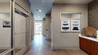 This contemporary commercial office space is thoughtfully designed to balance functionality with modern aesthetics. The single-floor layout features polished concrete flooring throughout, lending an industrial-chic character that ties the entire suite together cohesively.

The space is accessed through a bright, welcoming corridor with recessed lighting and clean grey walls, leading naturally into the core work areas. Multiple private offices — identified as Office One and Office Two — are enclosed by full glass-panel partitions and doors, offering both visual transparency and acoustic separation. Each office is equipped with built-in workstations and cabinetry, designed for efficient daily use.

A standout feature of the suite is the dedicated support area, which showcases a striking textured metallic tile accent wall that adds dimension and visual interest. Rich warm-toned wood cabinetry with white countertops lines this area, providing ample storage and workspace for printing and administrative tasks. This same area also houses a beverage station complete with a coffee maker and water dispenser, supporting a comfortable and productive work environment.

Natural light filters in through a glass-paneled front entrance, while interior clerestory windows between spaces maintain an open and connected feel. The cohesive grey palette, contemporary artwork, and polished surfaces throughout give this office suite a professional yet inviting ambiance.