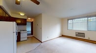 This single-floor unit offers a practical and straightforward layout across several distinct rooms. The open living area forms the heart of the space, featuring light-colored carpeting, neutral cream walls, and a ceiling fan with integrated lighting. A window unit air conditioner and baseboard heating provide year-round climate control. The living area transitions directly into the compact kitchen, which is equipped with dark wood cabinetry, light countertops, a gas range, a full-size refrigerator, a double sink, and an over-range microwave and exhaust hood. Tile flooring distinguishes the kitchen from the carpeted living spaces. Two bedrooms offer comfortable, quiet retreats, each with plush carpeting, cream-painted walls, and windows that frame views of surrounding greenery, allowing ample natural light to fill the rooms. Baseboard heating runs along the perimeter of each bedroom. The bathroom is finished with tile flooring, a wood-framed vanity with under-sink storage, a mirrored medicine cabinet, tiled shower-tub surround, and warm overhead lighting. Throughout the unit, a consistent neutral color palette and functional finishes create a clean, livable environment.