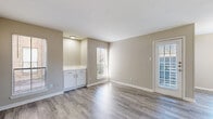 This single-floor space is designed with a clean, neutral aesthetic that creates a bright and cohesive atmosphere throughout. The main living area features wood-look plank flooring in a warm gray tone, complemented by soft beige walls and crisp white trim. A built-in cabinet with white shaker-style doors and a white countertop adds a functional accent to the living space. Multiple windows fitted with horizontal blinds allow generous natural light to fill the room, while a front entry door provides direct access to the exterior.

The kitchen is outfitted with white shaker cabinetry, light quartz-style countertops, and stainless steel appliances including a refrigerator and an electric range with an overhead ventilation hood, offering a practical and modern cooking environment.

The bedrooms are finished with plush carpet and feature neutral walls. At least one bedroom includes a walk-in closet with built-in shelving. A dedicated dining area benefits from a corner window arrangement that frames outdoor views and enhances the sense of openness.

The bathroom includes a white vanity with ample cabinet storage, a white countertop with an integrated sink, brushed nickel fixtures, a large mirror, and a tub-shower combination with tile surround, all set on wood-look tile flooring.