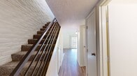 This two-floor space is defined by its striking architectural character, most notably the white-painted brick walls that appear consistently throughout, lending a cohesive and textured aesthetic across every room. The main floor hosts the living room, which stands out as the centerpiece of the space, boasting soaring vaulted ceilings with exposed beams, abundant natural light streaming through high clerestory windows and a sliding glass door, and a classic black fireplace set against a white brick surround. Wood-look flooring runs throughout the main level, adding warmth and continuity. The combined kitchen and living area features dark wood cabinetry, granite-style laminate countertops, white appliances including a range and dishwasher, and the same white brick backsplash that ties the space together. A carpeted staircase with metal railings leads to the upper floor, which offers a carpeted living area with a pass-through window overlooking the lower level, along with two closets equipped with built-in shelving. Two bathrooms serve the space — one featuring a granite-topped vanity and a full tub-shower combination, the other a half-bath with matching finishes. Textured ceilings complete the interior throughout.