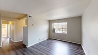 This single-floor residence offers a straightforward and functional layout designed to maximize everyday comfort. The living area features smooth gray wood-look laminate flooring that adds a clean, modern touch, complemented by neutral-toned walls and a large window fitted with horizontal blinds that invites natural light and outdoor views. A wall-mounted air conditioning unit provides additional climate control in this space.

The kitchen is compact yet practical, equipped with white cabinetry offering ample upper and lower storage, laminate countertops, a stainless steel sink, and a white gas range with oven. A refrigerator is also included, making the kitchen move-in ready.

The bedrooms are carpeted in warm beige tones, providing a soft and comfortable underfoot feel. Multiple windows dressed with horizontal blinds allow generous natural light to fill these rooms, creating a bright and airy atmosphere. The bedrooms offer versatile space suitable for various furniture arrangements.

Throughout the residence, the design maintains a clean, neutral palette with white trim and light gray walls, lending a cohesive and understated aesthetic. The single-level layout ensures easy navigation between spaces, making this a practical and well-organized home suited for a range of occupants.