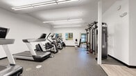 This fitness center is thoughtfully designed to support a full range of exercise routines across two distinct zones. The main cardio area features bright white walls, durable speckled rubber flooring, and long linear overhead lighting that keeps the space well-lit and energizing. This section is stocked with a row of treadmills, elliptical trainers, and stair climbers, alongside a comprehensive cable and strength machine system. Locker storage and posted safety guidelines add a practical, professional touch, while a dark-framed entry door provides clear room delineation.

The secondary exercise room offers a warmer atmosphere with soft yellow walls, light wood-style flooring, and large windows that invite natural light and outdoor views. This area is equipped with spin bikes, stability balls, an adjustable workout bench, a flat-screen television, and a rack of colorful dumbbells — making it ideal for group-style or functional training workouts. Decorative artwork featuring nature scenes enhances the calming ambiance. Together, the two areas create a comprehensive, amenity-rich fitness environment suited for both cardio endurance and strength-focused training.