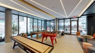 This contemporary amenity space is designed to encourage both active recreation and relaxed socializing. The game room is a standout feature, outfitted with a full-size shuffleboard table, a foosball table in bold red, and a billiards table set against a backdrop of floor-to-ceiling windows. Exposed concrete columns and linear LED lighting embedded in a tiered ceiling give the room an industrial-modern character, while abundant natural light and direct sightlines to the outdoor terrace create an open, energetic atmosphere.

Adjacent to the game room, a lounge area offers a more intimate setting furnished with vibrant orange swivel chairs, colorful modular ottomans, and small accent tables arranged for conversation and tabletop gaming. The same wall of black-framed glass partitions continues here, maintaining a seamless visual connection to the landscaped exterior terrace.

A dedicated media nook rounds out the space, featuring a built-in dark cabinetry unit with an integrated flat-screen display, accented by a distinctive wall clad in layered terracotta tile rolls that adds warmth and texture. Throughout, the design balances bold color, refined materials, and a strong indoor-outdoor flow, making this a versatile and visually engaging communal destination.