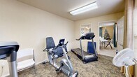 Located at 700 Cottage Park Cir in Guthrie, Oklahoma, this single-room fitness facility offers a dedicated space for cardiovascular exercise and general wellness. The room features warm, neutral-toned walls and patterned carpet flooring that adds a comfortable, welcoming feel underfoot. Overhead fluorescent lighting ensures the space is well-lit throughout, supplemented by natural light from windows dressed with blinds and curtain panels along one wall.

The fitness room is equipped with multiple cardio machines, including two treadmills and a recumbent stationary bike, providing a range of low- and high-impact workout options. A wall-mounted flat-screen television offers entertainment during exercise sessions, while a standing floor fan provides additional air circulation. A small set of hand weights is also present for light strength training.

Decorative wall art adds a personal touch to the otherwise utilitarian space. A pass-through window connects the fitness room to an adjacent common area, providing visual openness and a sense of connection to the broader facility. A standard door offers private access to and from the room. The overall layout is efficient and functional, making practical use of the available footprint to deliver a well-rounded amenity for residents of the Cottage Park community.