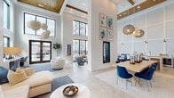 Located at 1139 Bragg Way in Myrtle Beach, SC, this thoughtfully designed community clubhouse offers a bright, open atmosphere defined by soaring double-height ceilings, expansive black-framed windows, and a seamless flow between distinct functional zones. The main lounge area commands attention with its dramatic volume, wood-accented ceiling beams, and woven pendant light fixtures that cast a warm glow over the space. A sleek, floor-to-ceiling linear fireplace serves as a striking architectural centerpiece, flanked by curated coastal artwork in soft blues and greens. The lounge is furnished with plush, modern seating arrangements set atop layered area rugs, creating an inviting gathering environment. Large glass doors open to outdoor views of palm trees and community amenities, seamlessly connecting the interior to the surroundings. A dedicated workspace area features built-in desks with computer stations, bathed in natural light streaming through two-story windows. The coffee and beverage bar is a refined nook finished with patterned tile, marble-look countertops, gray cabinetry with brass hardware, a wine refrigerator, and a professional espresso machine. Elegant arched doorways with rich wood trim connect the various areas, adding classic architectural character. Throughout, light-toned tile flooring, crisp white walls, and carefully selected furnishings contribute to a polished, coastal-modern ambiance.