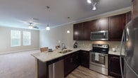 This single-floor residence offers a thoughtfully designed open-concept layout that seamlessly connects the kitchen and living areas. The kitchen is a standout feature, showcasing rich dark espresso cabinetry, light granite countertops, a peninsula with bar seating illuminated by elegant pendant lights, and a full suite of stainless steel appliances including a refrigerator, electric range, microwave, and dishwasher. Track lighting and crown molding add a polished finish to the space, while wood-look flooring grounds the kitchen and transitions into the carpeted living area. The living room is generously sized, featuring crown molding, a ceiling fan, and an arched doorway that adds architectural character. Natural light filters through multiple windows fitted with white blinds. The bedrooms are comfortable and well-lit, each with plush carpeting, ceiling fans, and large windows that bring in ample daylight. The bathroom is equipped with a tub-shower combination and a granite-topped vanity with dark cabinetry consistent with the kitchen's design language. A dedicated laundry room with a full-size side-by-side washer and dryer and overhead shelving provides added convenience. Overall, the space presents a cohesive, neutral palette with quality finishes throughout.