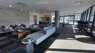 This contemporary communal lounge is designed with a sleek, open-plan layout that seamlessly integrates multiple functional zones into one cohesive space. The interior is anchored by a generous seating area furnished with modular gray sofas arranged around a striking acrylic side table, all set atop a geometric area rug. A cylindrical concrete column adds an industrial architectural accent while helping to define the zones within the open floor plan.

Toward the rear, a well-appointed kitchen area features pendant lighting suspended from the ceiling, a dark countertop bar with seating, and a bold patterned tile backsplash that serves as a focal point. Adjacent to the kitchen, a dining area offers round tables paired with modern curved-back chairs, ideal for casual gatherings or collaborative use.

Floor-to-ceiling glazing along the perimeter floods the space with abundant natural light, framing views of the surrounding urban cityscape. The exposed concrete wall panels and smooth white ceilings create a refined contrast, lending the space a polished yet industrial character. Dark tile flooring throughout ties the design together, contributing to the space's modern and sophisticated ambiance. The overall atmosphere is bright, airy, and thoughtfully curated for both comfort and functionality.