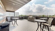 This contemporary rooftop terrace in Iowa City, Iowa, offers an inviting open-air retreat designed for socializing and relaxation. The spacious deck is finished with light gray composite decking that complements the modern aesthetic throughout the space. A built-in stainless steel gas grill station serves as a central amenity, providing a dedicated outdoor cooking area ideal for gatherings. Multiple high-top bistro tables with bar-height chairs are distributed across the terrace, creating several distinct seating zones for groups of varying sizes. A partially covered section features a striking accent wall composed of mixed-tone horizontal wood planks in warm browns, cool grays, and natural tones, adding a bold architectural focal point to the space. Recessed ceiling lighting beneath the covered overhang ensures usability during evening hours. The terrace is enclosed by low-profile perimeter barriers that preserve sweeping views of the Iowa City skyline, neighboring rooftops, and lush tree canopies extending into the distance. The open sky exposure fills the space with abundant natural light throughout the day. The overall design balances contemporary materials with functional outdoor living, making this rooftop terrace a well-appointed communal space.