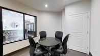 This single-floor space presents a clean, contemporary room designed for intimate meetings or dining. At its center sits a round, dark granite-topped table supported by a sleek chrome pedestal base, surrounded by four matching black leather chairs that lend a polished, professional feel. The walls are finished in crisp white, contributing to a bright and minimalist aesthetic, while dark hardwood-style flooring grounds the space with a contrasting richness.

A recessed ceiling light provides focused overhead illumination, keeping the atmosphere well-lit without being harsh. A standout architectural feature is the interior window along one wall, framed in black, which looks through into an adjacent room where a chandelier and additional furnishings are visible. This interior glazing adds depth and visual interest, creating a sense of openness between spaces.

A solid white paneled door with a chrome lever handle provides access, maintaining the room's clean, modern character. The combination of neutral tones, sleek furnishings, and thoughtful design details makes this a functional and stylish space suited for small gatherings or focused conversation.