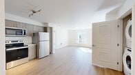 Located on SE Eastgate Way in Bellevue, WA, this two-bedroom, three-bathroom residence offers a thoughtfully designed single-floor layout. Light-toned hardwood-style flooring runs throughout the main living areas, contributing to a bright, cohesive aesthetic complemented by white walls and abundant natural light from well-placed windows.

The open-concept living room flows seamlessly from the kitchen, which features gray wood-grain cabinetry extending to the ceiling, white countertops, a double sink, and stainless steel appliances including a dishwasher, range, and microwave. A track lighting fixture illuminates the kitchen area effectively.

The primary bedroom is a well-proportioned space with a large window providing natural light and an adjacent walk-in closet with built-in shelving. The second bedroom features soft carpeting and direct access to the main living area. Both rooms include individual thermostats for climate control.

Three bathrooms are finished consistently, each featuring wood-grain vanity cabinetry, white countertops, and hardwood-style flooring. Tub-shower combinations are included in the full bathrooms. A stacked in-unit washer and dryer provides added convenience. The overall design is clean, modern, and functional.