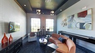 This contemporary lounge space makes a strong first impression with its striking design and thoughtfully curated interiors. The room's most distinctive architectural feature is its deep navy tray ceiling, which is adorned with a combination of recessed lighting and modern pendant fixtures that cast a warm, ambient glow throughout the space. Large, gridded windows line the walls, flooding the interior with natural light and offering views of surrounding greenery.

The lounge is furnished with a comfortable mix of seating options suited to both casual relaxation and informal gatherings. A curved, salmon-toned sectional sofa anchors the main seating area, complemented by a sculptural metallic coffee table and coordinating accent tables. A crisp white accent chair adds contrast, while navy throw pillows tie the color palette together cohesively.

Along the windowed wall, a high-top table with bar-height seating creates an additional casual nook, ideal for conversation or light work. A sleek, dark credenza runs along one wall, adding functional storage while contributing to the room's refined aesthetic. Decorative wall art, including a vibrant abstract canvas and a cluster of circular mirrored accents, add personality and visual interest. The deep-toned, botanical-patterned carpet grounds the space and reinforces its sophisticated, modern ambiance.