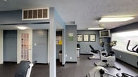 Carlton Apartments includes a well-appointed fitness center designed to meet the exercise needs of its residents. The space is finished with cool blue-gray walls and durable speckled flooring, creating a clean and functional environment suited for regular workouts. The room is equipped with cardio machines, including a stationary bike and an elliptical trainer, positioned for comfortable use. A wall-mounted flat-screen television is installed at an elevated position, allowing residents to stay entertained during their workout sessions. Structural columns divide the space subtly, adding architectural character without disrupting the open flow of the room. Overhead fluorescent lighting ensures the area is well-lit throughout. A dedicated information board near the entry displays notices and guidelines for residents. A waste bin and water dispenser are conveniently placed within the space for ease of use. The textured ceiling and climate control vents reflect a practical, utility-focused design. Overall, the fitness center at Carlton Apartments provides a straightforward and accessible amenity space that supports an active lifestyle for all residents.