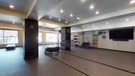 This dedicated fitness studio offers a thoughtfully designed space for functional and high-intensity training. The room features durable rubberized flooring throughout, providing a safe and supportive surface for a wide range of workouts. Recessed lighting set into a textured ceiling illuminates the space evenly, creating a bright and energizing environment.

At the center of the studio, structural columns wrapped in branded padding serve a dual purpose — protecting users during dynamic movements while reinforcing the facility's identity. Battle ropes and a heavy tire are positioned on the open floor, catering to strength and conditioning exercises. Along one wall, a well-organized rack holds an assortment of medicine balls in various sizes, alongside a workout sled and additional training equipment. A wall-mounted storage system displays colorful resistance bands and accessories, keeping essentials accessible and neatly arranged.

Large windows along one side of the room allow natural light to filter in, complementing the overhead lighting. A floor-to-ceiling mirror on one wall expands the sense of space and allows users to monitor their form during training sessions. The overall layout is open and spacious, offering ample room for group workouts or individual training circuits.
