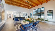 This expansive single-floor amenity space is defined by its striking architectural design, blending modern aesthetics with warm, natural elements. Soaring ceilings lined with rich wood beams and exposed timber rafters create a dramatic sense of height and openness throughout the main lounge area. A cascading pendant chandelier with elongated black fixtures serves as a bold focal point, illuminating the open-concept gathering space below.

The main lounge is furnished with contemporary blue armchairs, a plush green velvet sofa, and a clean-lined white coffee table, all anchored by a layered striped area rug. Large black-framed floor-to-ceiling windows flood the interior with abundant natural light and offer views of an outdoor pool and terrace area, with direct access via full-height glass doors.

The fully equipped kitchen features a dramatic geometric-patterned backsplash with gold accents, quartz waterfall countertops, dark cabinetry, stainless steel appliances including a professional-grade range and oversized refrigerator, and pendant lighting over a substantial island with bar seating.

Multiple dining areas include a casual table with green velvet chairs near the pool-view windows, a larger communal dining setup with bold pendant lighting, and a dedicated private conference or meeting room outfitted with a long wood table, quilted leather chairs, a wall-mounted display, and curated artwork—ideal for professional gatherings.