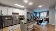 This thoughtfully designed single-floor residence offers a seamless open-concept layout that connects the kitchen, dining, and living areas into one cohesive, functional space. The kitchen is a standout feature, showcasing two-tone cabinetry with crisp white upper cabinets and gray lower cabinets, dark granite countertops, a decorative stone-tile backsplash, black appliances, and a central island with bar seating — ideal for casual dining or entertaining. Wood-look flooring flows throughout the main living areas, enhancing the warm, modern aesthetic. The living room is anchored by a bold teal accent wall and a sliding glass door that opens onto a private balcony, filling the space with natural light and outdoor views of lush greenery. A distinct entryway, also featuring deep teal walls, provides a welcoming transition into the home. Both bedrooms are well-appointed, offering generous natural light through large windows dressed with gray curtains, wood-look flooring, and a calm, neutral palette. The bathroom features a full soaking tub and shower combination with clean white tile surround and contrasting dark accent walls. Throughout the residence, a cohesive color scheme of teal, gray, and warm wood tones creates a modern yet inviting ambiance.