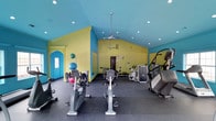 This fitness center offers a lively and energetic atmosphere, distinguished by its bold color scheme of bright yellow-green walls contrasted against a vivid turquoise ceiling. The vaulted ceiling adds a sense of openness and height to the space, while recessed lighting and a ceiling fan ensure the environment remains well-lit and comfortable during workouts. The flooring consists of durable rubber matting, practical for exercise and easy maintenance. The facility is well-equipped with a range of cardio machines, including stationary bikes and an elliptical trainer, positioned toward the front of the space for easy access. Toward the rear, a cable weight machine and additional strength training equipment provide options for resistance-based workouts. Exercise balls stored along the wall offer versatility for functional fitness routines. A turquoise-trimmed arched doorway adds a decorative architectural detail consistent with the space's colorful design. Overall, the fitness center combines a cheerful, motivating aesthetic with a practical layout, providing users with a comprehensive range of equipment in a compact, well-organized environment.