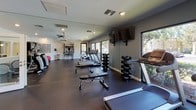 Located at 5100 Quail Run Rd in Riverside, California, this single-floor fitness center is thoughtfully designed to support a wide range of workout routines. The space is divided into distinct workout zones, each equipped with professional-grade exercise equipment. A dedicated cardio area features multiple treadmills and an elliptical machine, positioned alongside large windows that provide an abundance of natural light and views of the surrounding greenery, creating an inviting atmosphere for endurance training. Full-length mirrors line the walls, visually expanding the space and allowing users to monitor their form during workouts. A wall-mounted flat-screen television enhances the cardio experience. The strength training zone is well-stocked with a full dumbbell rack, adjustable weight benches, and a multi-function cable machine suitable for a variety of resistance exercises. A functional training area houses a wall-mounted rack equipped with kettlebells, suspension straps, and additional free weights, alongside a stationary bike and a stability ball. The space benefits from recessed lighting, ceiling fans, and rubber flooring throughout, ensuring both comfort and safety. A welcome sign and posted fitness center rules contribute to the organized, community-oriented environment. Direct access to the outdoor area is available through a glass-panel door.
