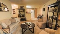 This single-floor space showcases an open-concept layout that seamlessly connects the living, dining, and kitchen areas, creating a functional and cohesive flow throughout. The living area is carpeted and comfortably sized, accommodating a cushioned sofa, an armchair, and a round dark-wood coffee table as its centerpiece. A freestanding bookshelf along the wall provides both storage and decorative display space, adding character to the room.

The kitchen is visible from the living area through a pass-through opening, featuring a white refrigerator and warm wood-toned cabinetry. A fluorescent ceiling fixture illuminates the kitchen workspace efficiently. Adjacent to the kitchen, a compact dining area is furnished with a small table and chairs, suitable for intimate meals.

Throughout the space, warm yellow-toned walls contribute to an inviting ambiance, complemented by soft carpeting underfoot. Decorative elements including framed wall art, candles, and accent pieces add a personalized touch. Ceiling vents suggest a central HVAC system for year-round climate control. The overall layout is practical and well-organized, making efficient use of the available space.