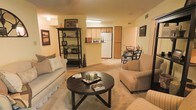 This single-floor space showcases an open-concept layout that seamlessly connects the living, dining, and kitchen areas, creating a functional and cohesive flow throughout. The living area is carpeted and comfortably sized, accommodating a cushioned sofa, an armchair, and a round dark-wood coffee table as its centerpiece. A freestanding bookshelf along the wall provides both storage and decorative display space, adding character to the room.

The kitchen is visible from the living area through a pass-through opening, featuring a white refrigerator and warm wood-toned cabinetry. A fluorescent ceiling fixture illuminates the kitchen workspace efficiently. Adjacent to the kitchen, a compact dining area is furnished with a small table and chairs, suitable for intimate meals.

Throughout the space, warm yellow-toned walls contribute to an inviting ambiance, complemented by soft carpeting underfoot. Decorative elements including framed wall art, candles, and accent pieces add a personalized touch. Ceiling vents suggest a central HVAC system for year-round climate control. The overall layout is practical and well-organized, making efficient use of the available space.